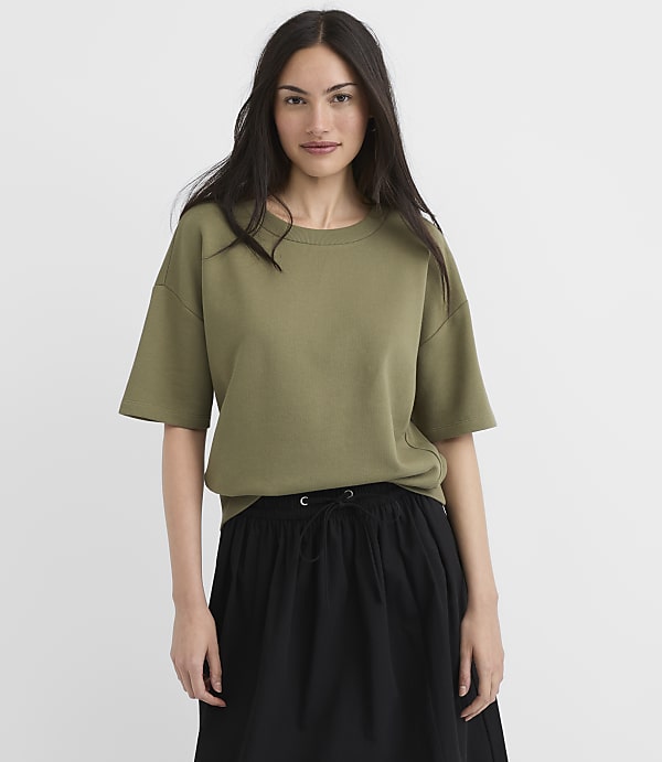 A woman in a green t-shirt and black skirt stands against a plain white background.