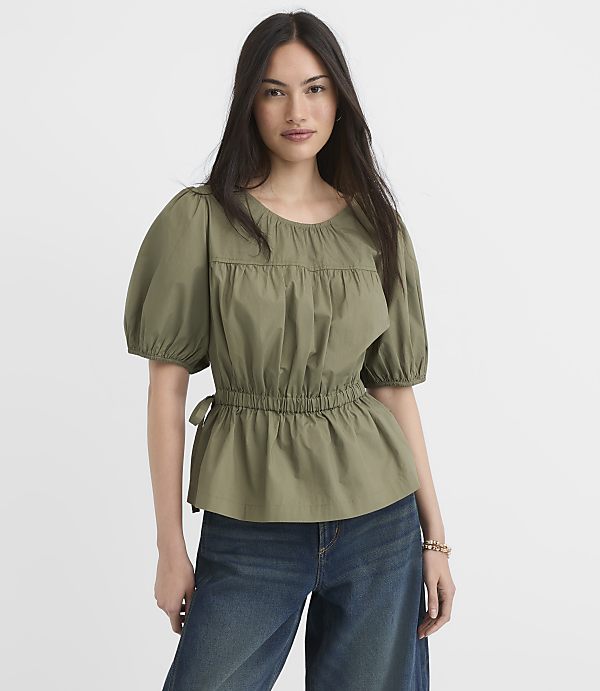 A woman in a green puff-sleeve top and blue jeans stands against a plain white background.