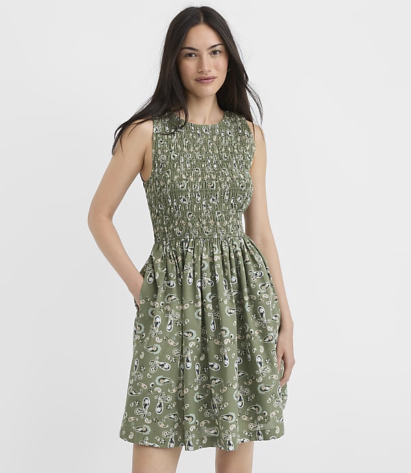A woman in a green paisley-print dress stands against a plain white background.