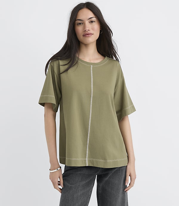 A woman in a green t-shirt and dark jeans stands against a plain gray background.