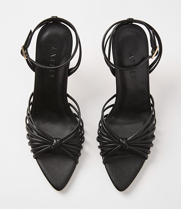 A pair of black strappy high-heeled sandals with knotted details in the foreground are set against a plain white background.