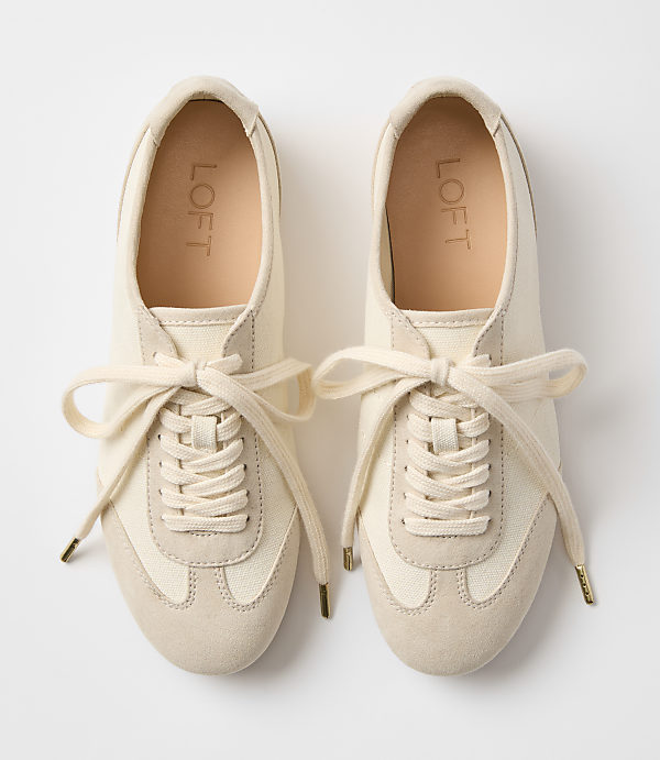 A pair of cream-colored canvas and suede sneakers with cream laces are laid out on a plain white background.