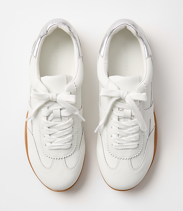 A pair of white leather sneakers with silver accents and gum soles rests on a plain white background.