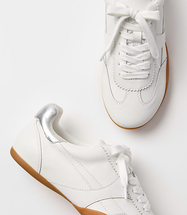 Two white leather sneakers with gum soles and silver heel accents rest on a plain white background.