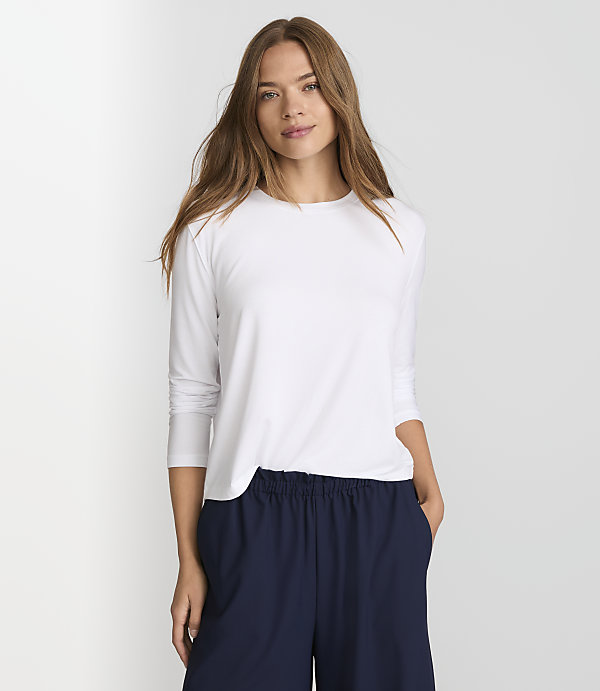 A woman in a white long-sleeved shirt and navy pants stands against a plain white background.