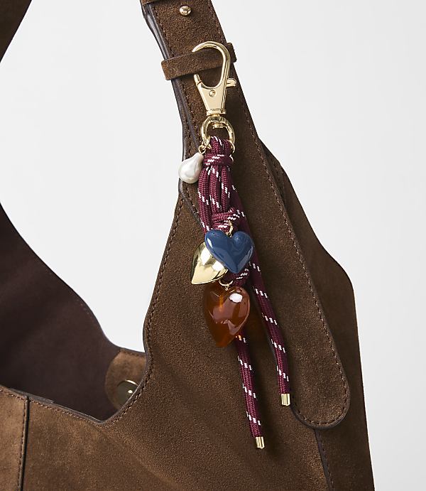A brown suede bag with a decorative charm featuring hearts and a pearl hangs in the foreground, against a plain white background.