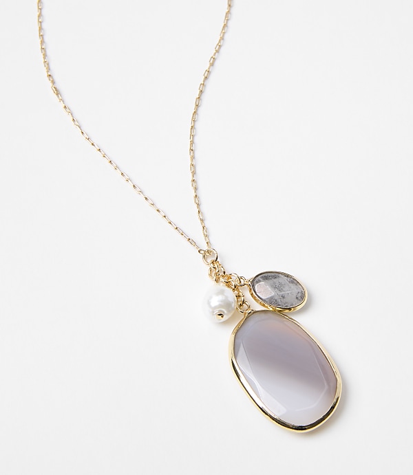 A delicate gold chain necklace with three charms, including a pearl and two polished stones, rests against a plain white background.