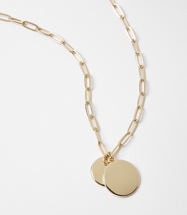 A gold paperclip chain necklace with two circular pendants rests on a plain white background.