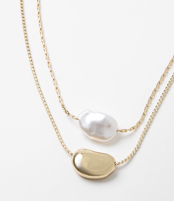Two delicate gold chains, one adorned with a lustrous baroque pearl and the other with a smooth, golden pebble, rest against a clean white background.