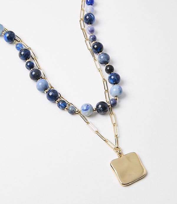 A layered necklace with blue beads and a gold square pendant rests on a plain white background.