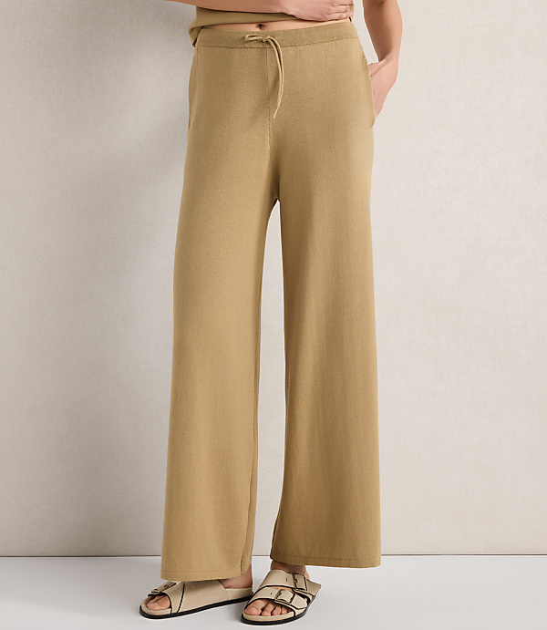 A person stands in tan wide-leg pants and sandals against a textured off-white background.