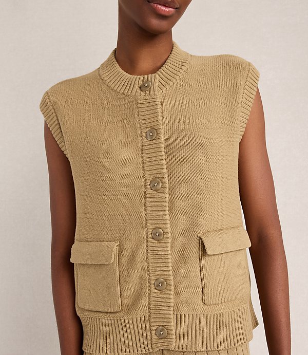 A close-up of a person wearing a tan knit vest with buttons and pockets is set against a plain, light-colored background.