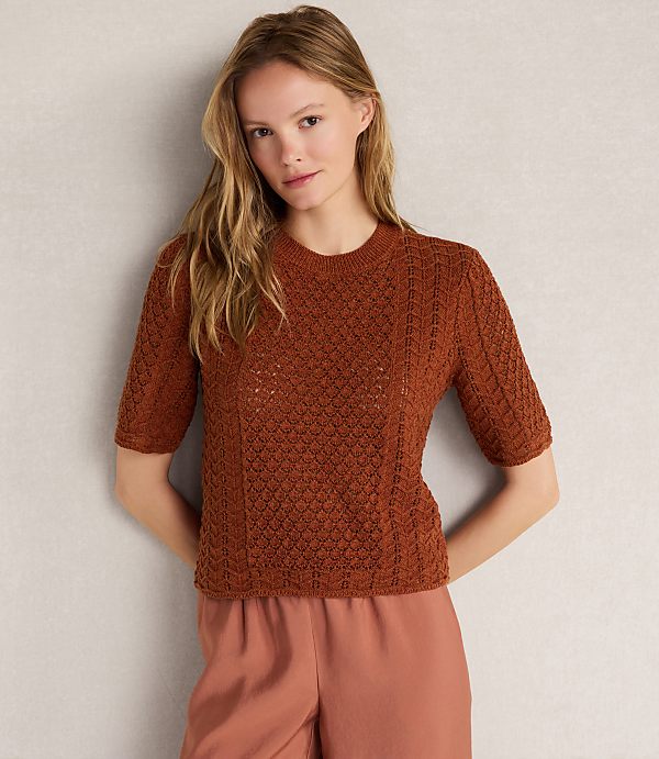 A woman in a brown knit top and pink pants stands against a textured gray background.