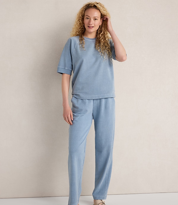 A young woman with curly blonde hair stands in a light blue loungewear set against a plain, light-colored background.