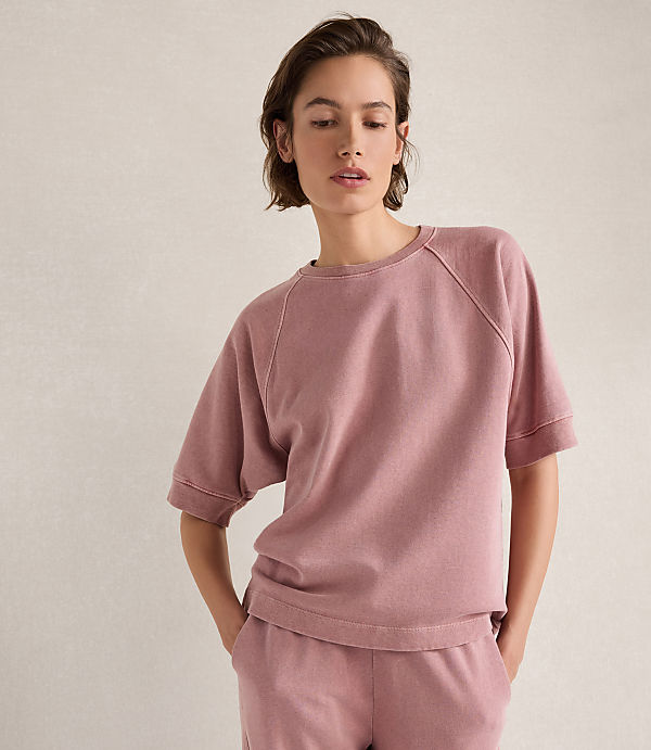 A woman in a pink sweatshirt and matching pants stands against a textured, light-colored wall.