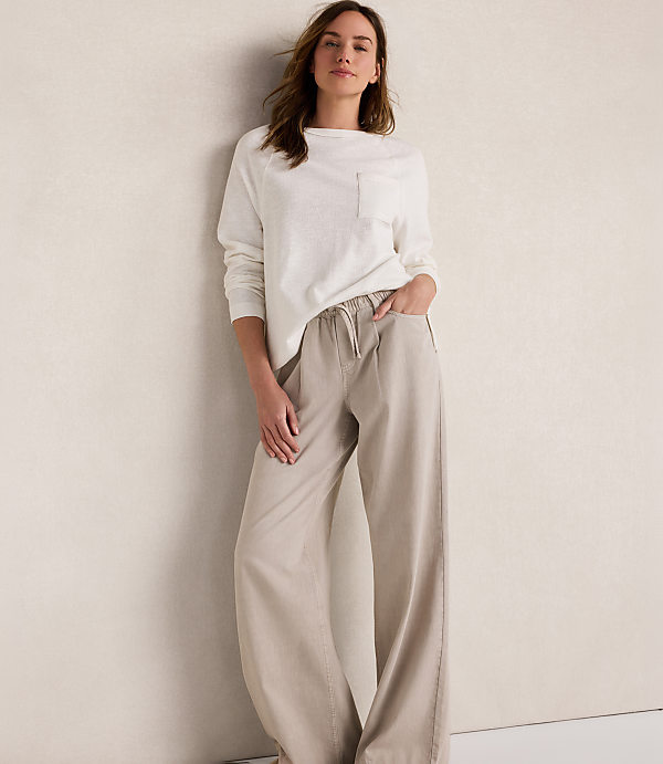 A woman in a white long-sleeved shirt and beige wide-leg pants stands against a textured cream wall.