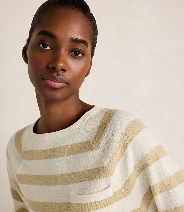 A woman with dark skin and short hair wears a cream and tan striped sweater with a pocket in the foreground, against a softly lit, textured background.