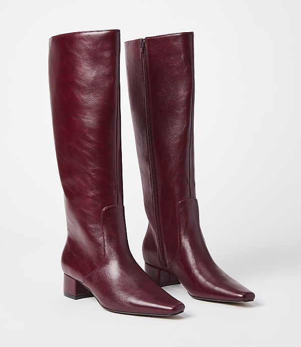 A pair of burgundy leather knee-high boots with block heels stand on a white surface against a plain white background.