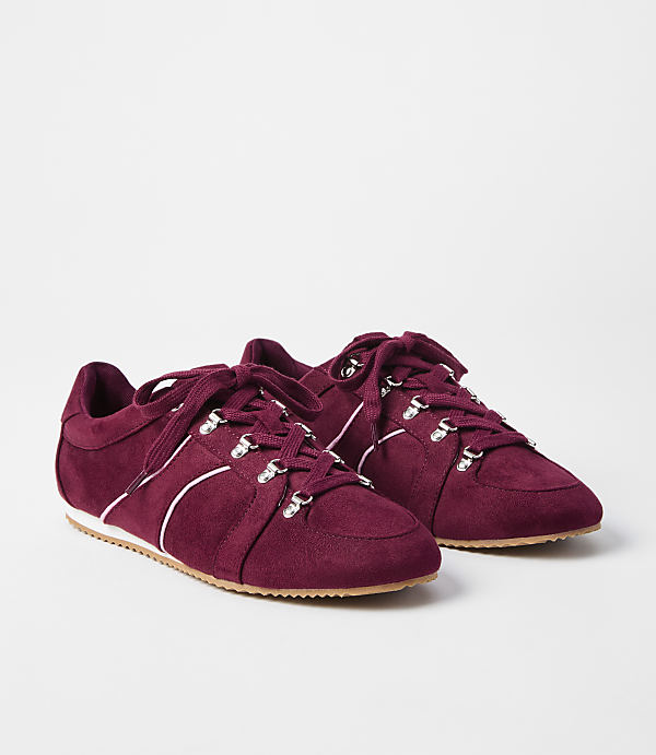 A pair of burgundy suede sneakers with white laces and silver eyelets rests on a white surface against a plain white background.