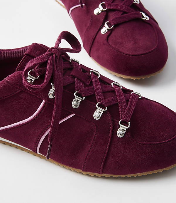 Burgundy suede sneakers with pink accents and silver eyelets are in the foreground, with another shoe blurred in the background.
