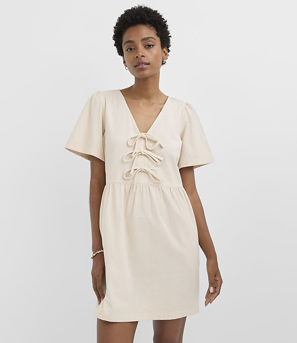 A woman in a cream-colored dress with flutter sleeves and a tie-front neckline stands against a plain white background.