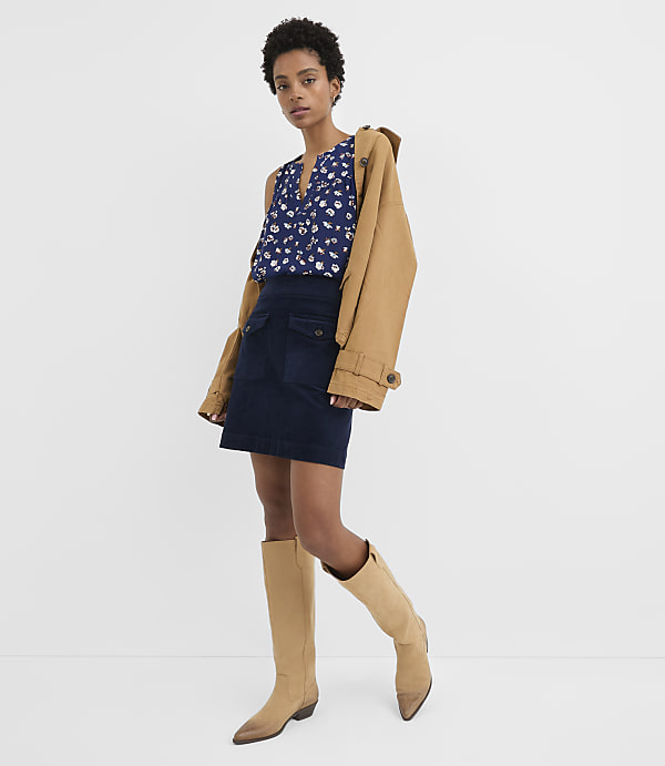 A woman in a floral top, denim skirt, and tan boots stands against a plain white background with a tan jacket draped over her shoulder.