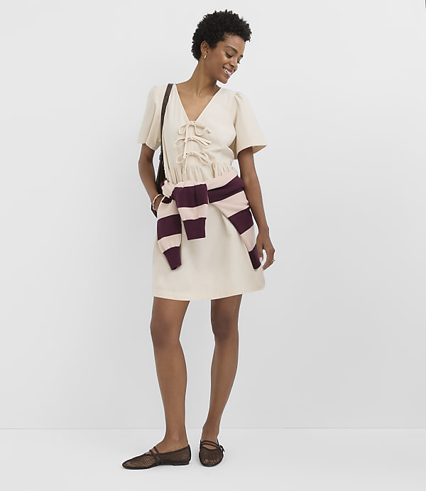 A woman in a cream dress and sheer flats stands against a plain white background, with a striped sweater tied around her waist.