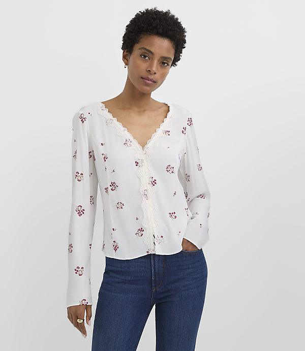 A woman in a white floral blouse and blue jeans stands against a plain white background.