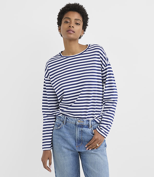 A woman in a striped long-sleeved shirt and jeans stands against a plain white background.