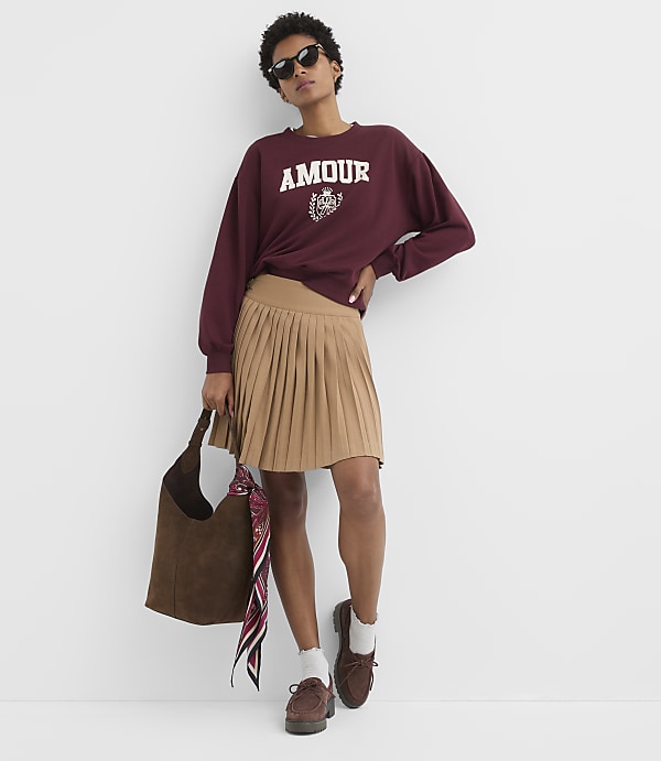 A woman in a burgundy sweatshirt and tan pleated skirt stands against a plain white background, holding a brown suede tote bag.