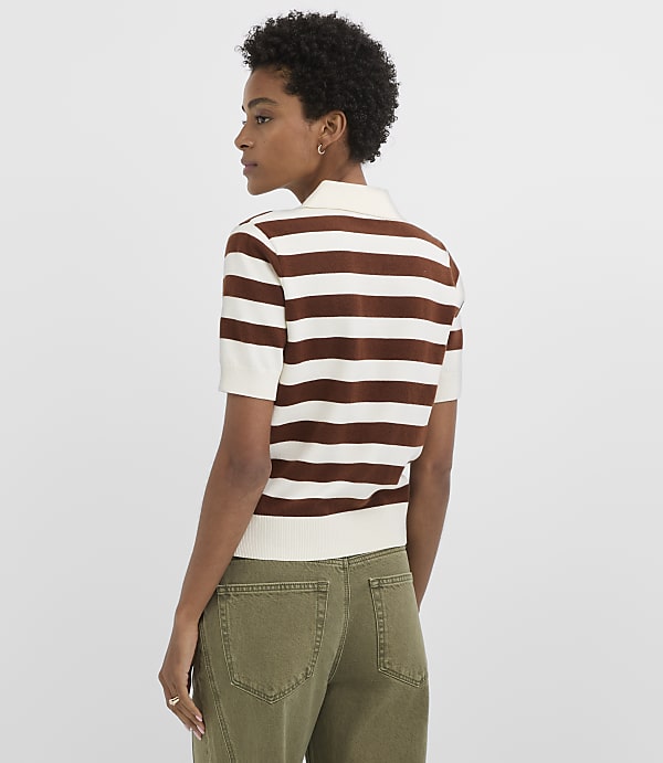 A woman in a brown and white striped shirt and green pants stands against a plain white background.