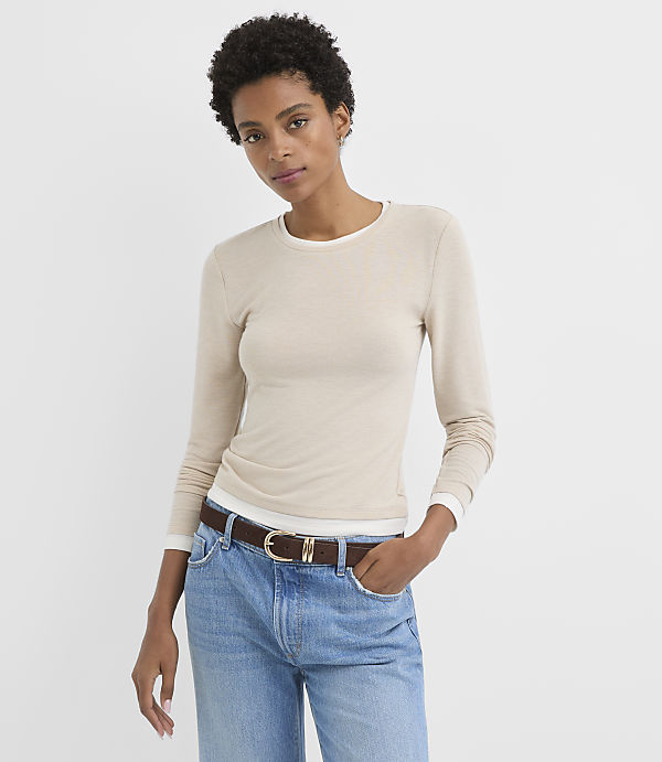 A woman in a beige long-sleeved shirt and blue jeans stands against a plain white background.