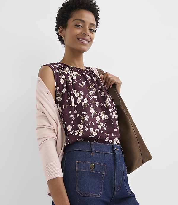 A woman in a floral top and denim pants smiles against a plain background, with a pink cardigan draped over her shoulder and a brown bag slung across her back.