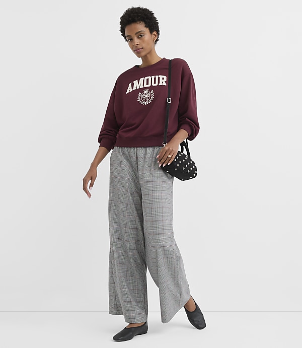 A woman in a maroon sweatshirt and plaid pants stands against a plain white background, holding a studded black purse.