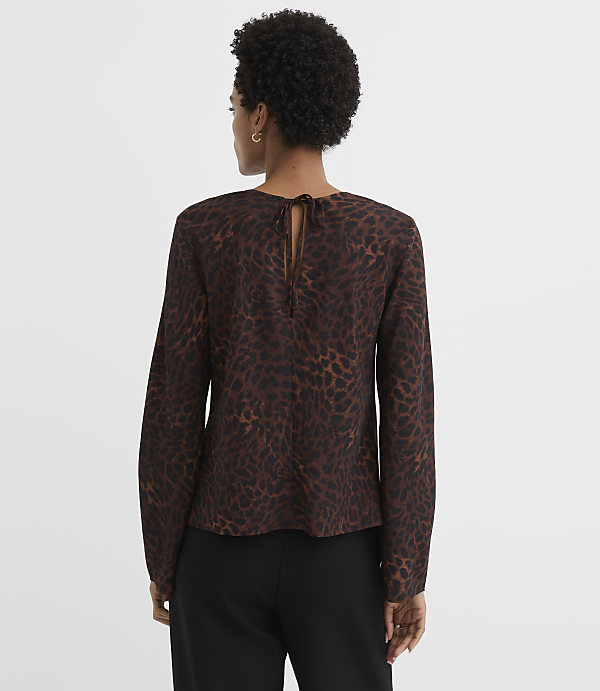 A woman in a brown leopard-print blouse with a tie at the back stands against a plain, light brown background.