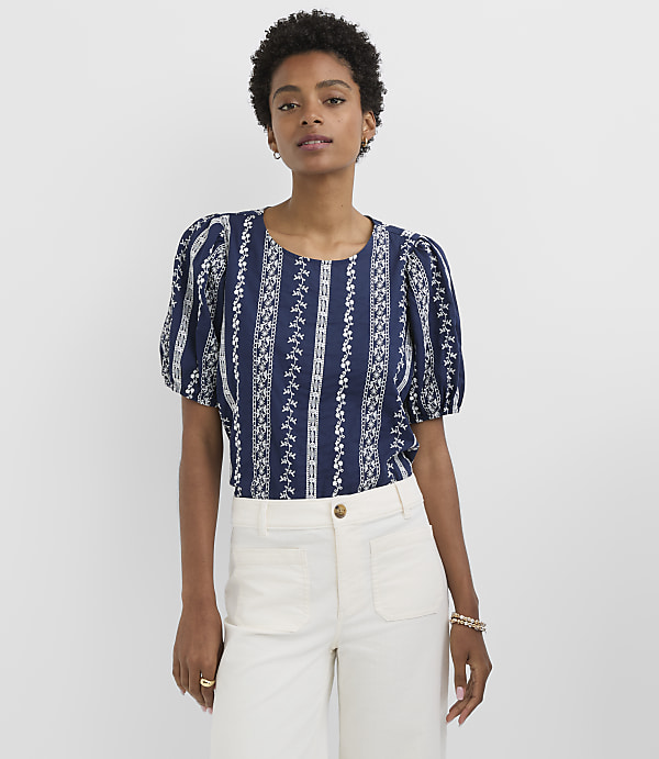 A woman in a patterned blue top and white pants stands against a plain white background.