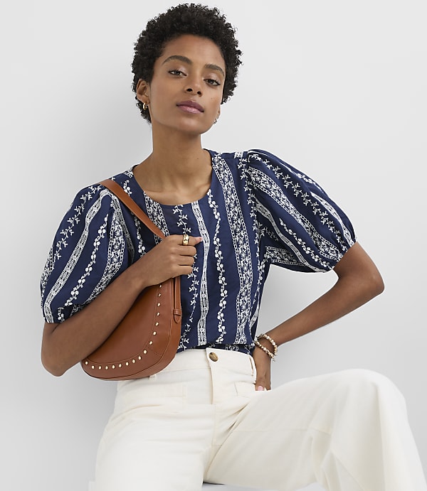 A woman in a patterned blue top and white pants poses with a brown saddlebag against a plain white background.