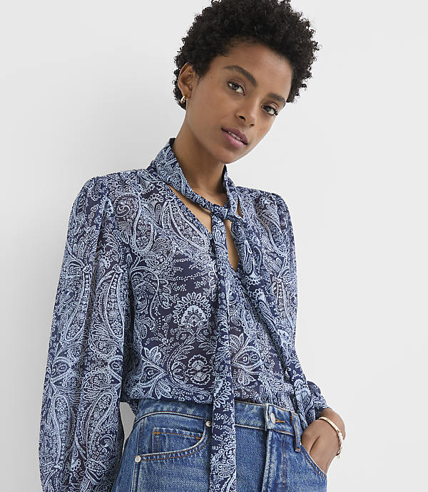 A woman in a blue paisley blouse and jeans stands against a plain white background.