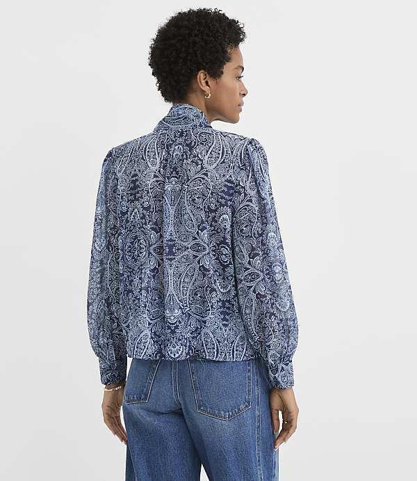 A woman in a blue paisley blouse and jeans stands against a plain white background.