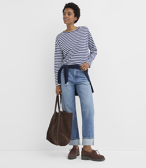 A woman in a striped shirt and cuffed jeans stands with a brown tote bag in front of a plain white background.