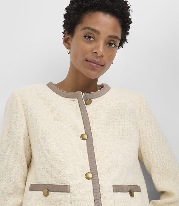 A woman in a cream-colored tweed jacket with brown trim and gold buttons stands against a plain gray background.