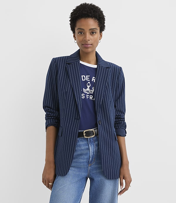 A woman in a pinstripe blazer and jeans stands against a plain white background.