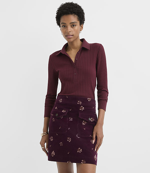 A woman in a burgundy ribbed polo shirt and a floral corduroy skirt stands against a plain white background.