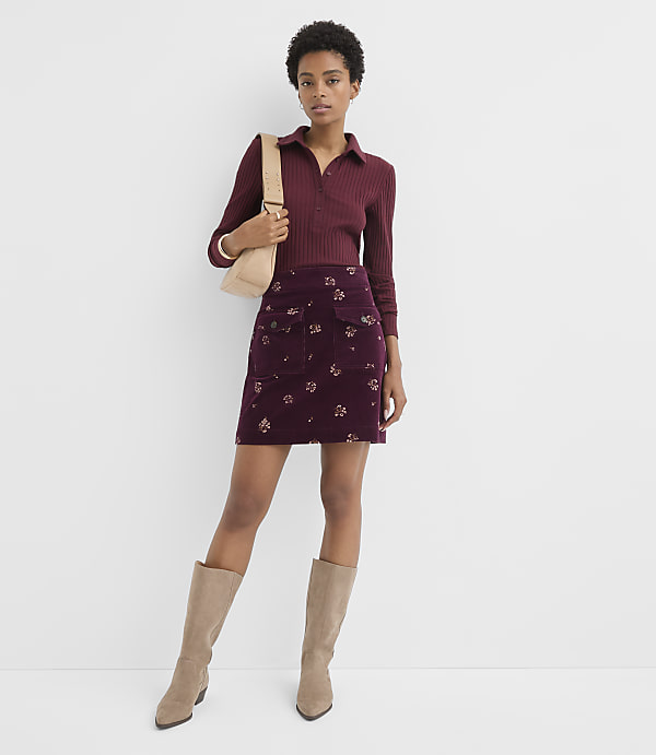 A woman in a burgundy ribbed polo shirt and a floral print mini skirt stands against a plain white background, accessorized with a beige crossbody bag and knee-high boots.