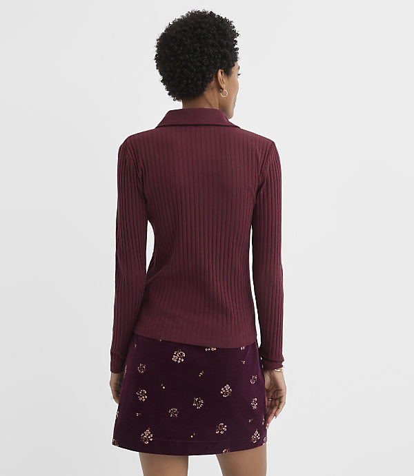 A woman in a burgundy ribbed turtleneck and a floral skirt stands against a plain white background.