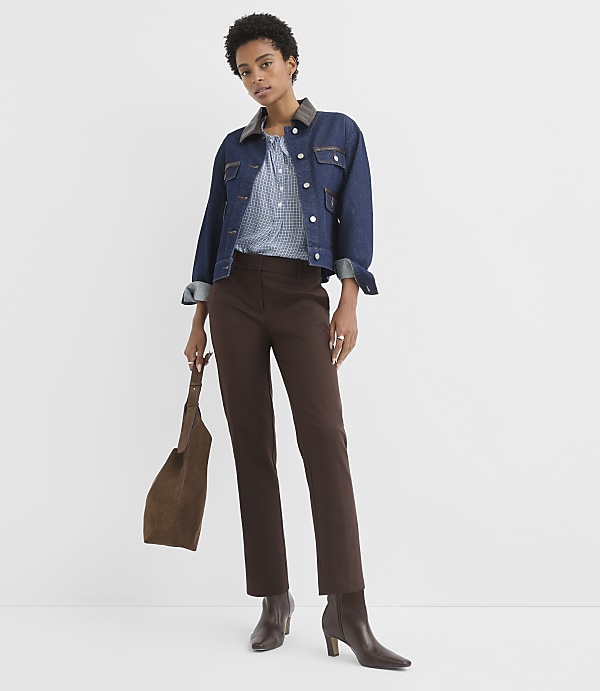 A woman in a denim jacket and brown pants stands against a plain white background, holding a brown suede purse.