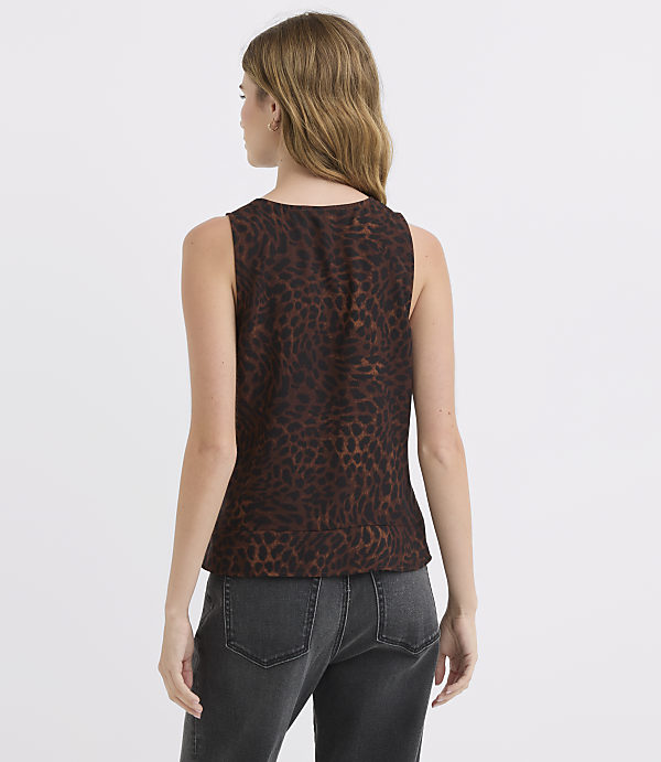 A woman wearing a brown leopard-print sleeveless top and dark jeans stands against a plain, light brown background.