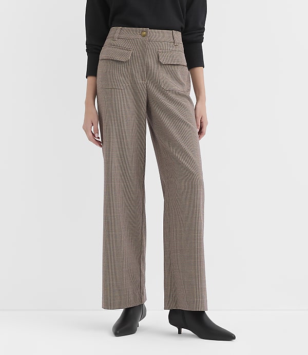 A person stands in front of a plain white background, wearing a black top and brown plaid wide-leg pants with black heeled boots.
