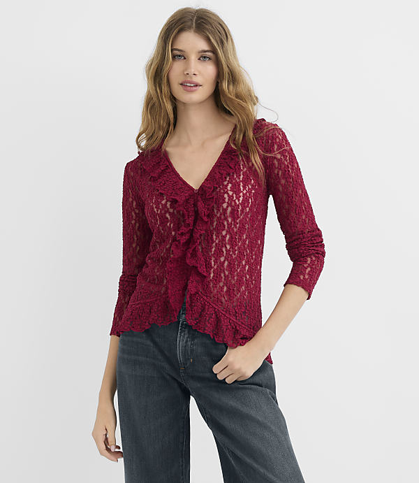 A woman in a red lace cardigan and dark jeans stands against a plain white background.