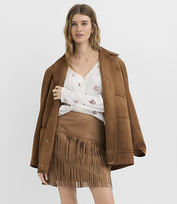 A woman in a brown suede jacket and fringed skirt stands against a plain white background.